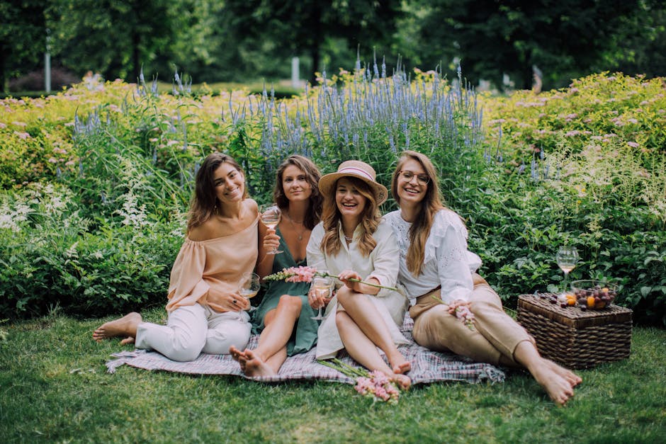 A group of four women sitting on a striped picnic blanket in a lush garden, surrounded by blooming flowers and greenery, enjoying a sunny day. They are smiling and holding glass glasses of white wine,