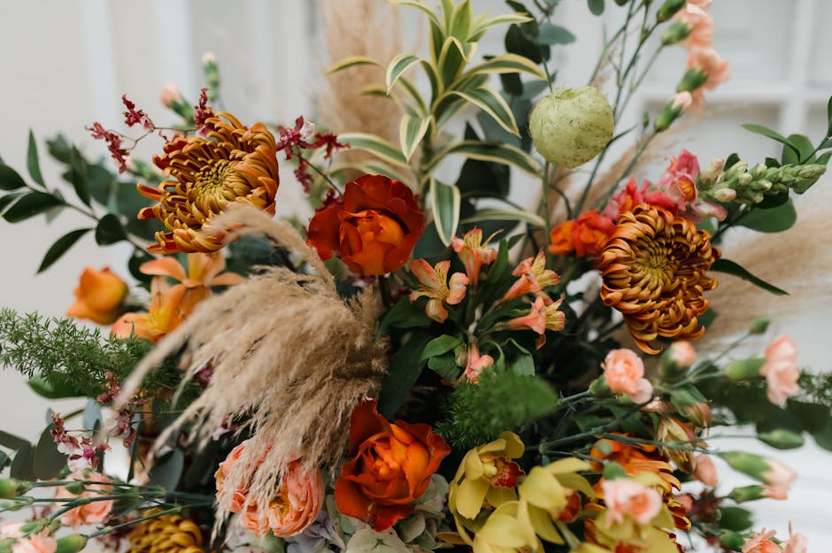 A close-up of a vibrant flower arrangement featuring orange and yellow roses, large variegated green and white leaves, and various smaller pink and peach blossoms. The bouquet includes textured pampas