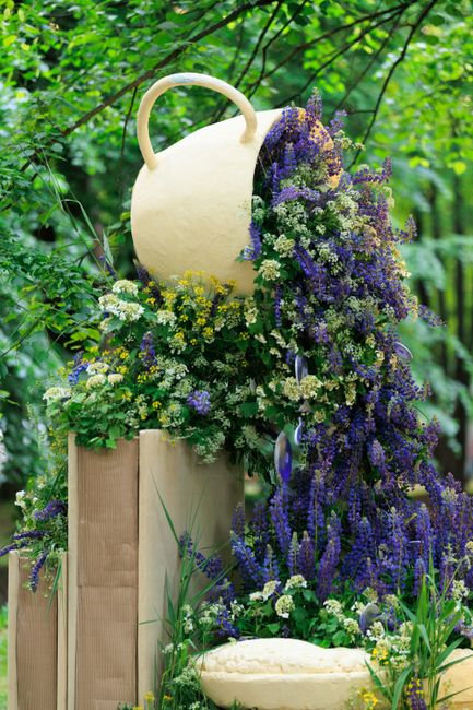A floral display featuring a large, cream-colored ceramic jug positioned on a wooden pedestal outdoors, surrounded by an arrangement of fresh flowers in shades of purple, white, and yellow. The flower