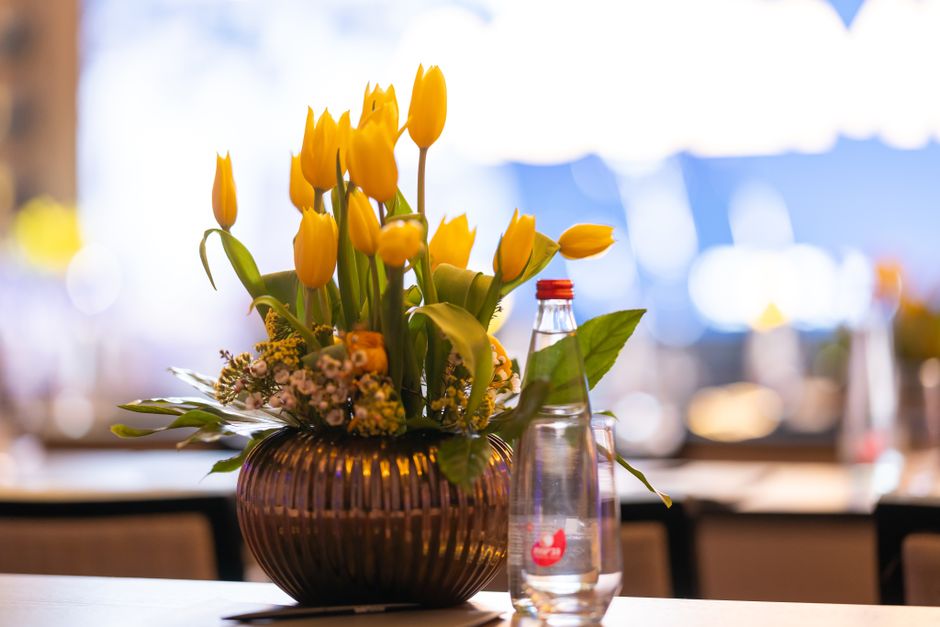 A floral arrangement featuring a bouquet of vibrant yellow tulips with closed and open petals, complemented by small pink and white filler flowers, all set in a ribbed, dark amber glass vase. The vase