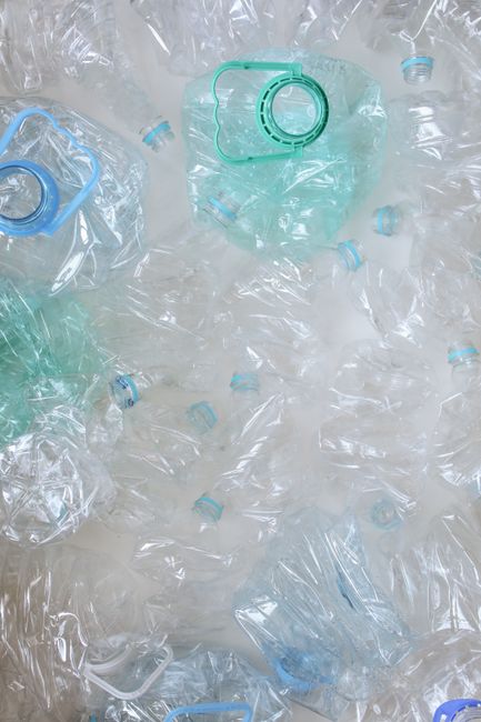 A collection of clear plastic water bottles and plastic bottle caps, some with attached tamper-evident rings, scattered across a light-colored surface. The bottles are empty, with a few containing res