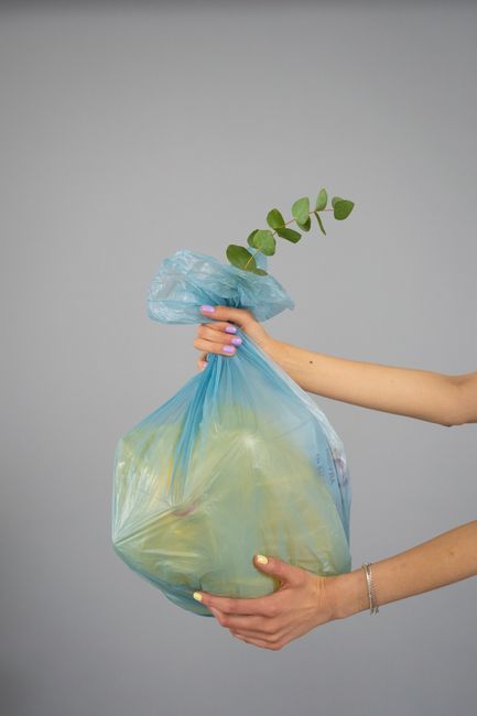 A person holds a large, transparent plastic bag containing fresh flowers and greenery, with green leaves and stems visible through the bag. The person’s hands and forearms are visible, with one hand