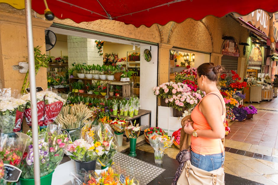 A woman in a peach sleeveless top and blue jeans stands on a sidewalk in Hampstead Village, holding a bag and examining a vibrant floral arrangement outside a florist shop. The shop, part of Florist H