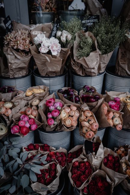 A display of fresh flowers arranged in grey metal buckets and wrapped in brown paper, featuring various types of roses in shades of pink, cream, peach, and red. Some bouquets include additional greene